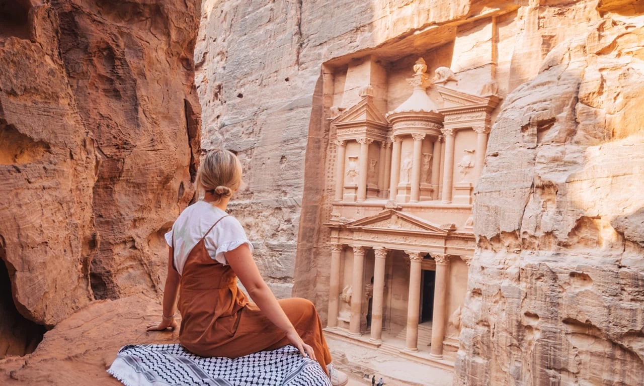 A tourist sitting in front of the Treasury in Petra, surrounded by sandstone cliffs.